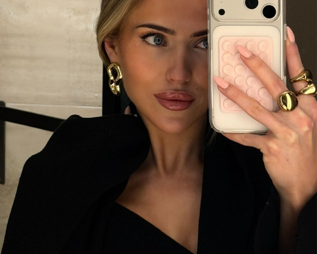 A women taking a selfie in the mirror. I. her hand she is holding her hand and has her three golden rings. In her ear she is wearing the golden Valeria earring. 