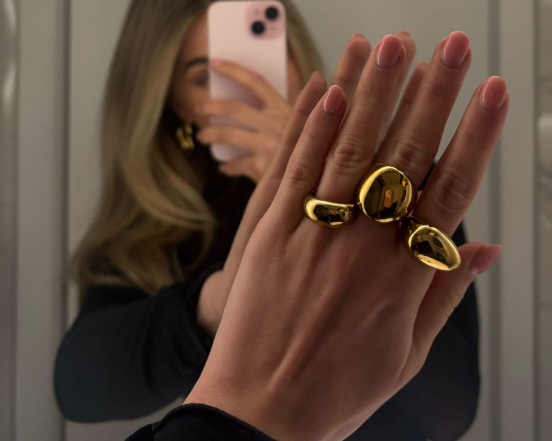 Photo in the mirror of a blonde women. She makes a picture with her phone of her hand with three golden rings. 