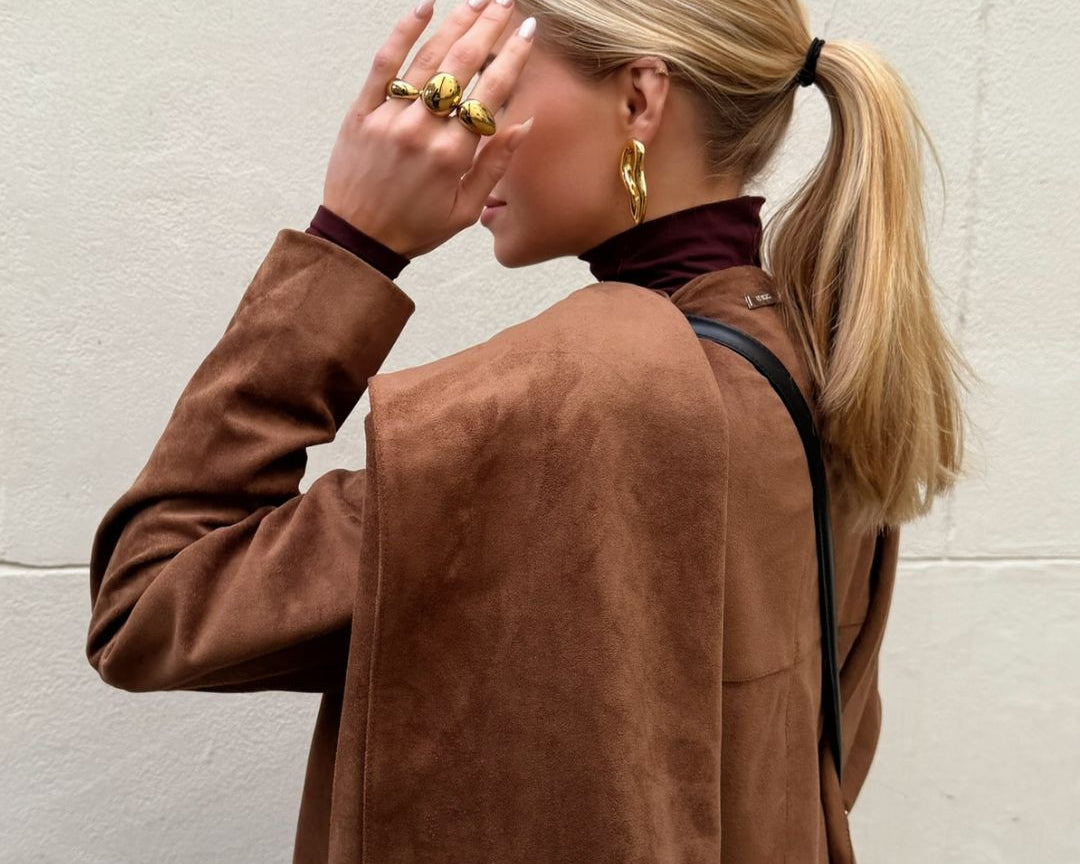 A blonde women on the street. She stands in front of a white wall. You can see the side of the upper body and head. She is holding her hand in front of her face wearing the three golden rings. She is also wearing the golden Valeria earring. 